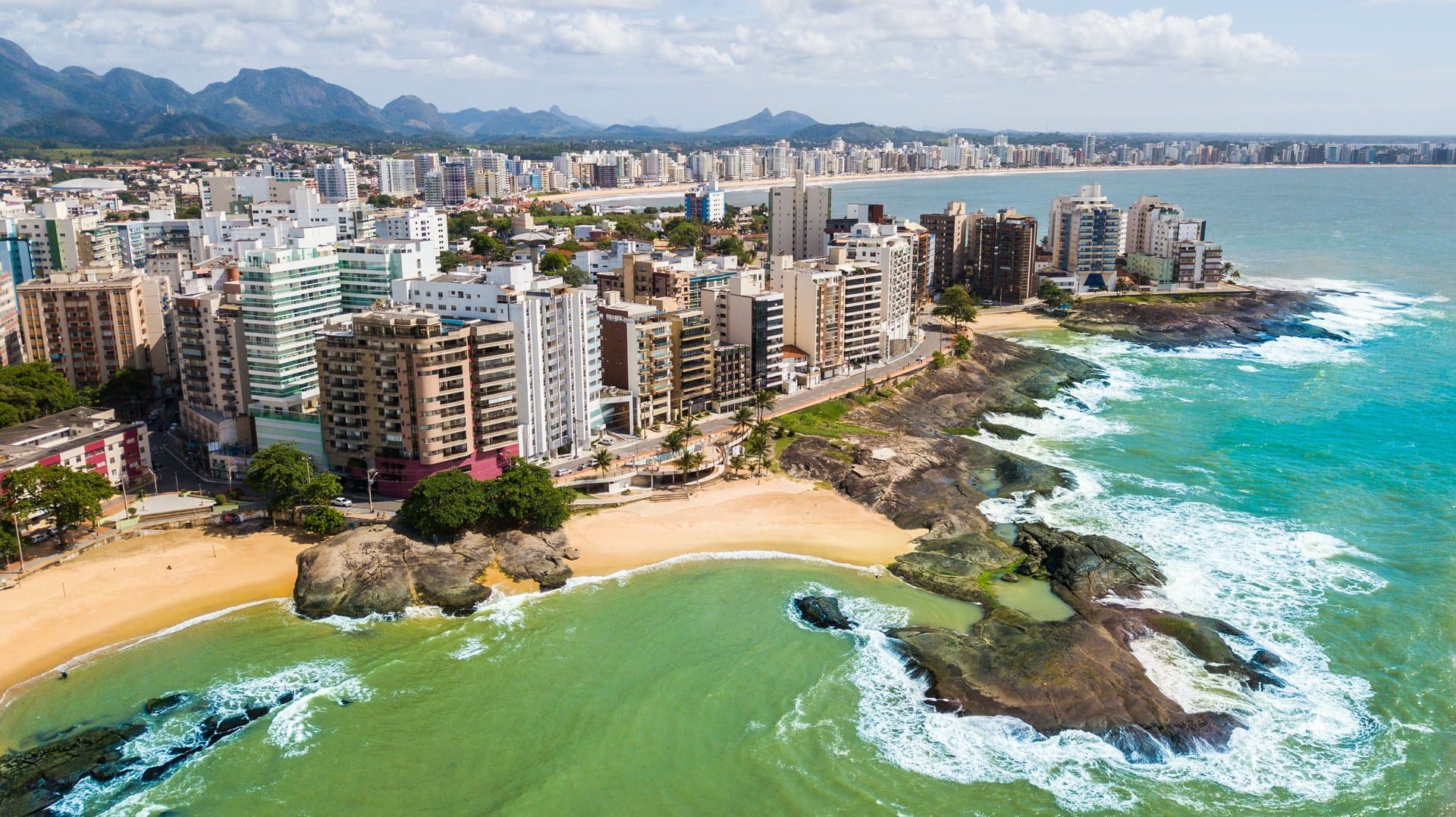 Vista aérea de Guarapari, com prainhas entre pedras escuras, prédios à beira-mar e a serra ao fundo